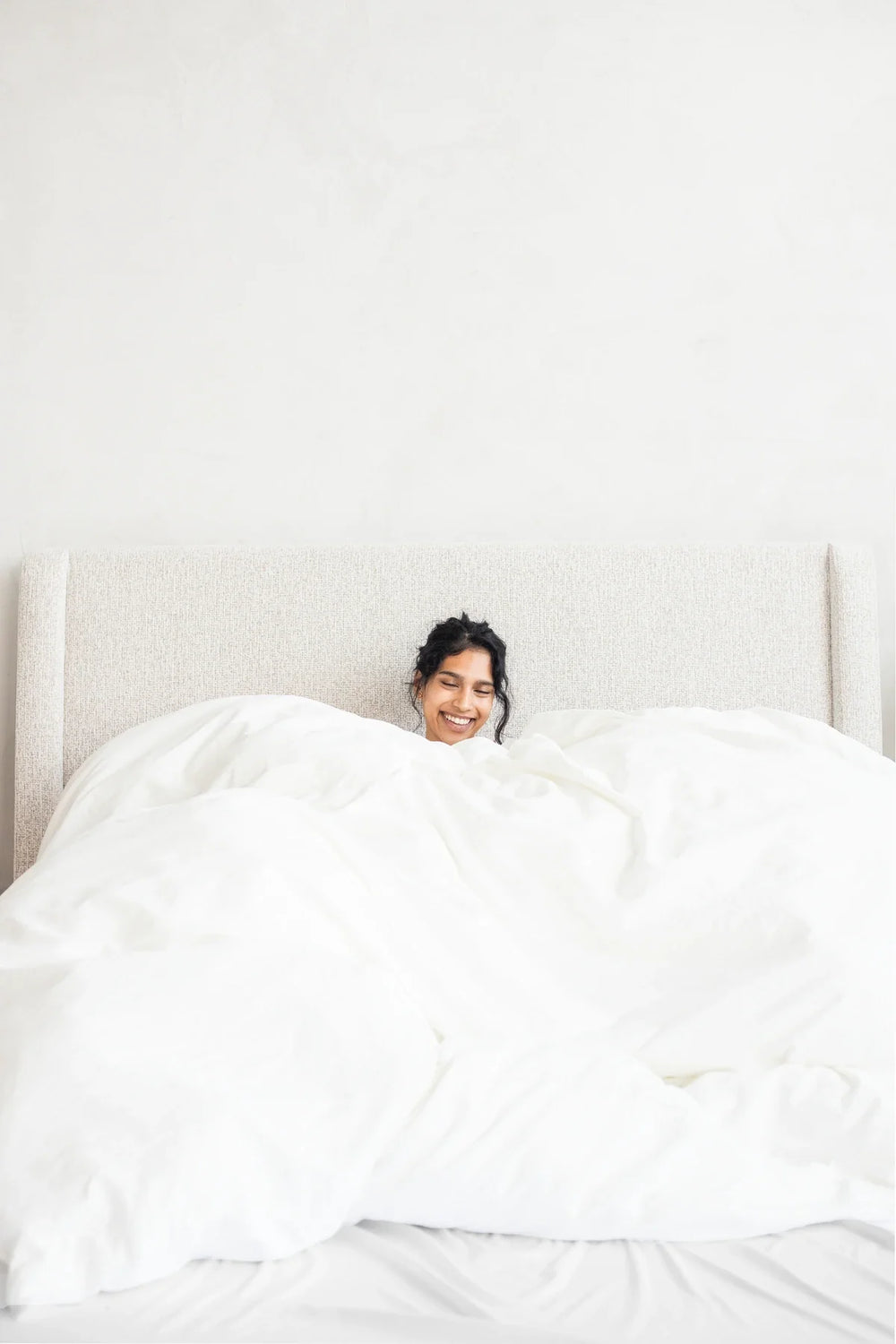 A woman smiling from beneath a pile of white duvet.